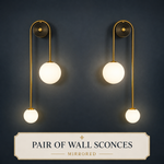 A pair of modern wall sconces with gold-finished metal arms and glowing white spherical glass shades, mounted on a dark navy wall. A cream label at the bottom reads 'PAIR OF WALL SCONCES MIRRORED'.