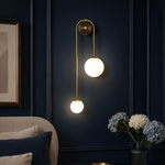 A modern wall sconce with two glowing white globe bulbs on a curved gold brass arm, mounted on a dark navy blue paneled wall above a bed in a styled bedroom.
