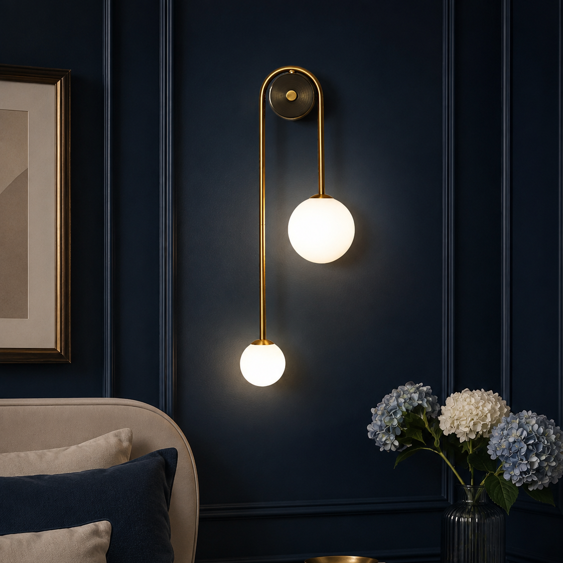 A modern wall sconce with two glowing white globe bulbs on a curved gold brass arm, mounted on a dark navy blue paneled wall above a bed in a styled bedroom.