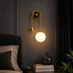 A modern wall sconce with a curved gold brass arm, a spherical gold finial, and a glowing white globe shade, mounted on a dark gray wall above a bedside table.