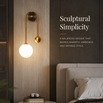 A modern wall-mounted sconce with a curved gold metal arm and a glowing white globe bulb, installed above a bedside table in a styled bedroom. Text on the wall reads, 'Sculptural Simplicity' with descriptive copy below.