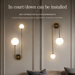 Two modern wall sconces with gold-toned curved metal arms and glowing white spherical bulbs mounted on a paneled gray wall above a sofa. Text at the top reads 'In court/down can be installed' with smaller installation information.